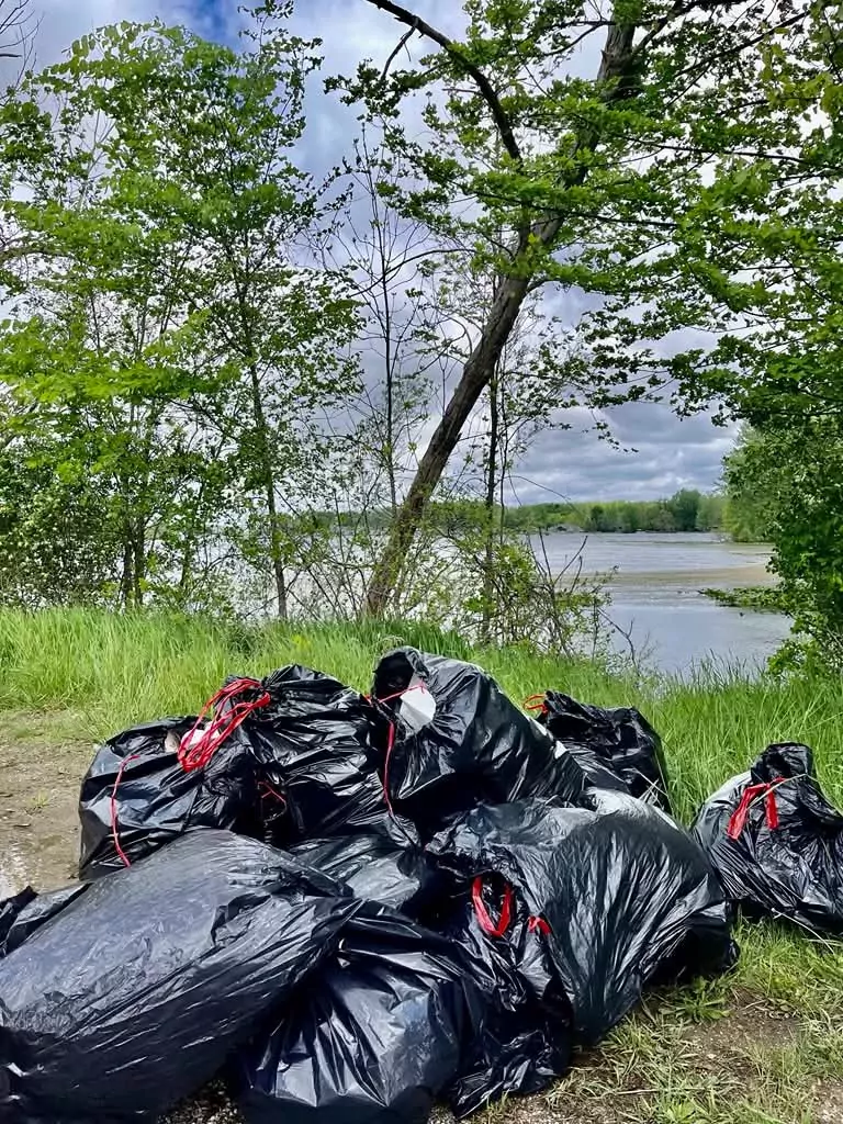 A pile of roughly a dozen full black trash bags on the grass at the top of a lake bank.