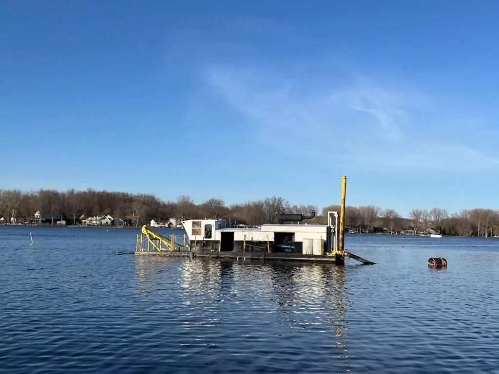 A flat dredging barge with a crane and a spud pole sitting on open water, houses along the far shore.