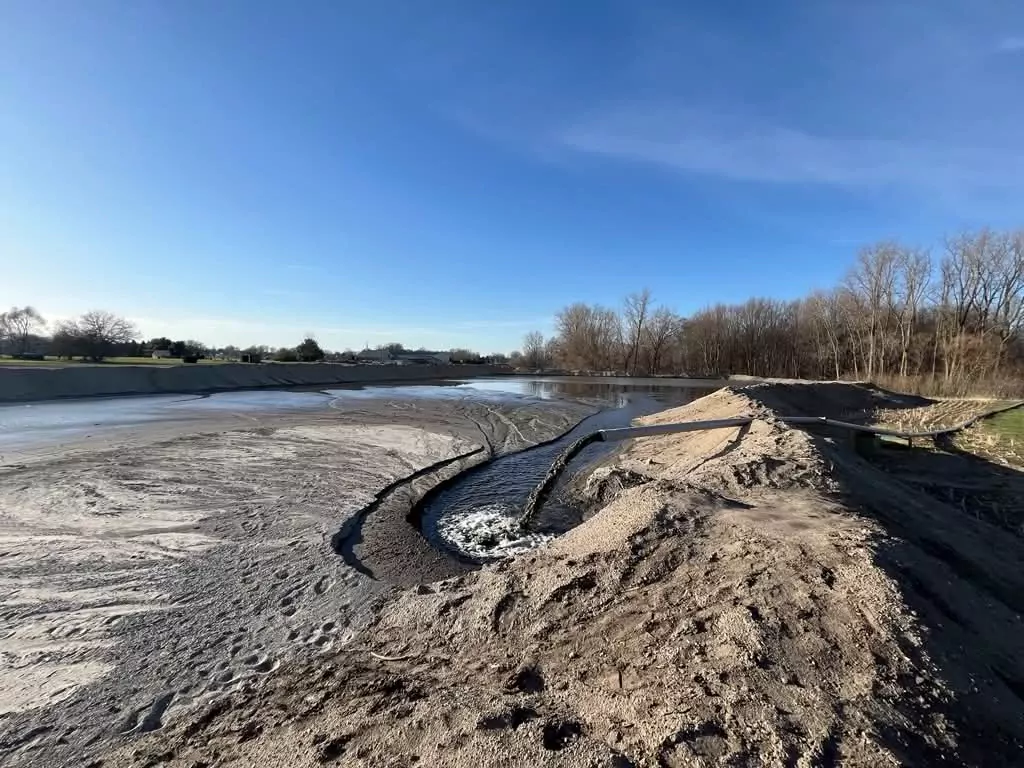A wide bare basin of grey sediment behind sand berms, with a discharge pipe running into standing water.