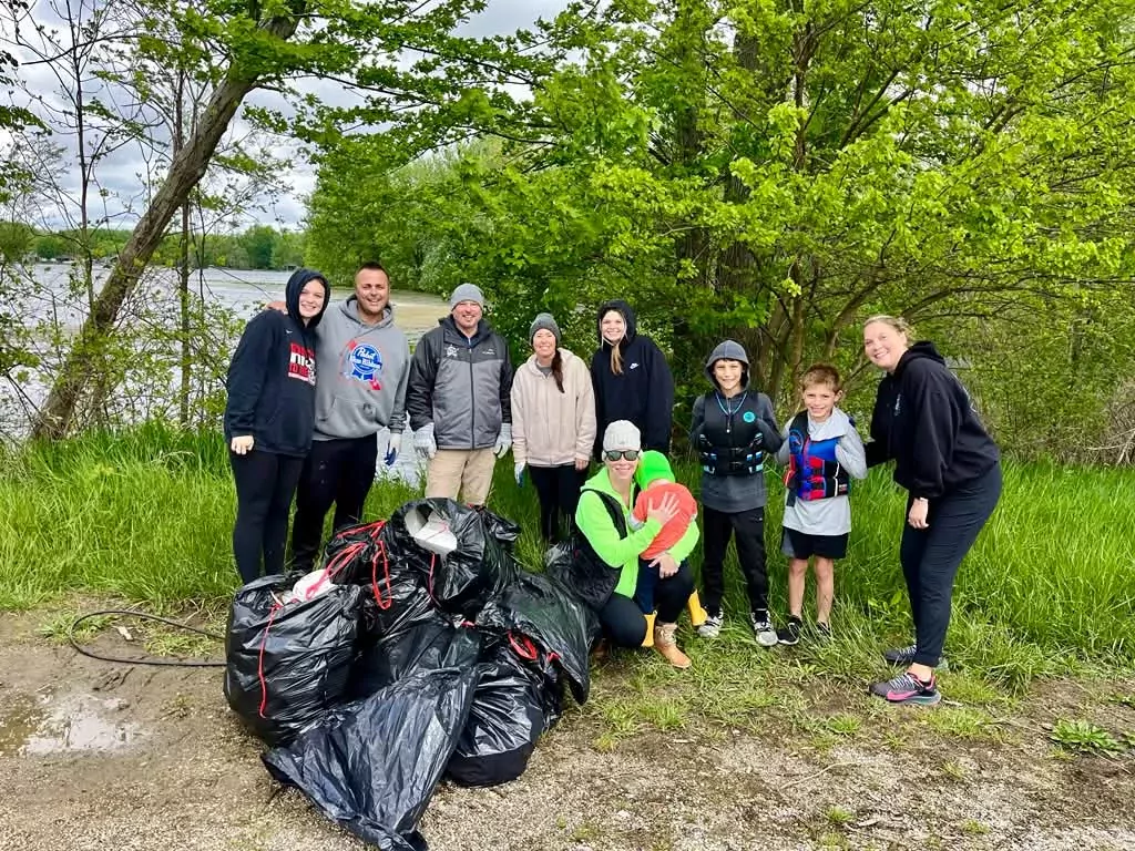About ten adults and children standing on a grassy bank beside the lake, next to a row of filled black trash bags.